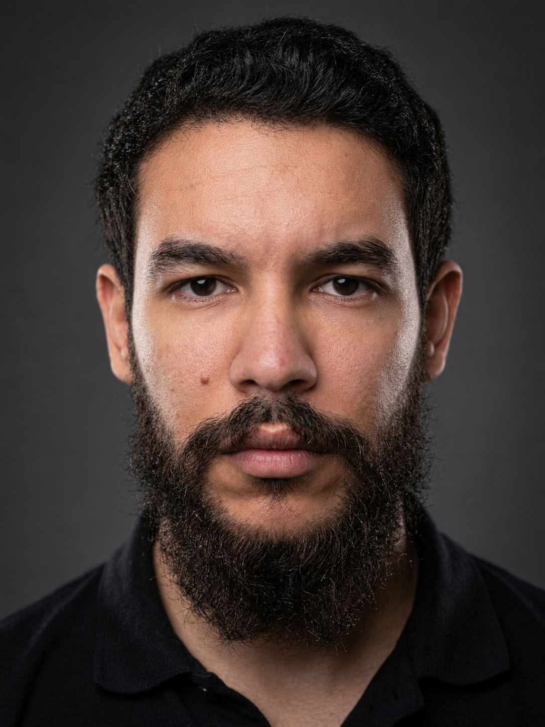 Portrait of Mehdi Ghalime: a man with short dark hair and a full dark beard, wearing a black polo shirt, looking straight at the camera against a dark grey background